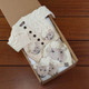 Sheply the Sheep Aran Cardigan Gift Set featuring cream-colored knitted baby cardigan with sheep designs, matching hat and mittens, arranged in an open cardboard box on a wooden surface. Sheply the Sheep Aran Cardigan Gift Set featuring cream-colored knitted baby cardigan with sheep designs, matching hat and mittens, arranged in an open cardboard box on a wooden surface.